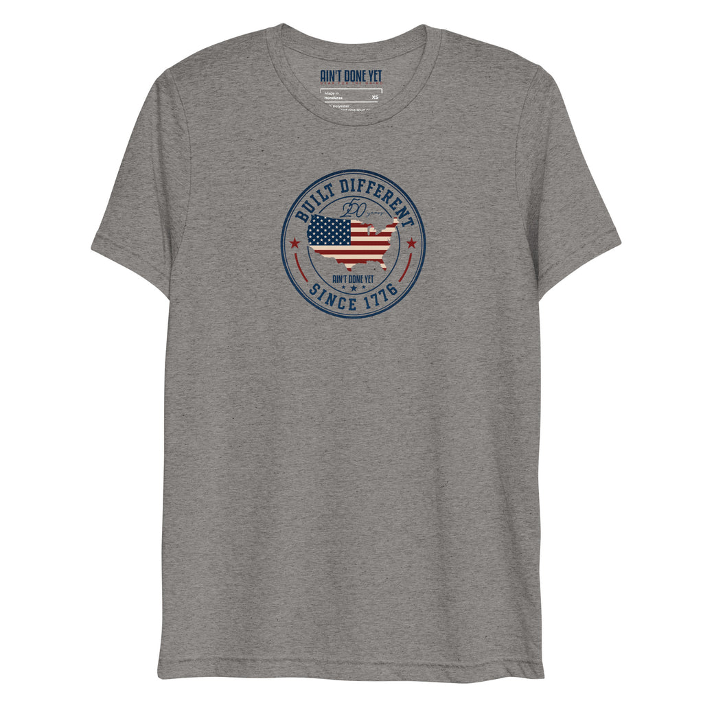 Built Different America 250 grey tri-blend t-shirt featuring patriotic circular emblem with American flag and 1776 design.