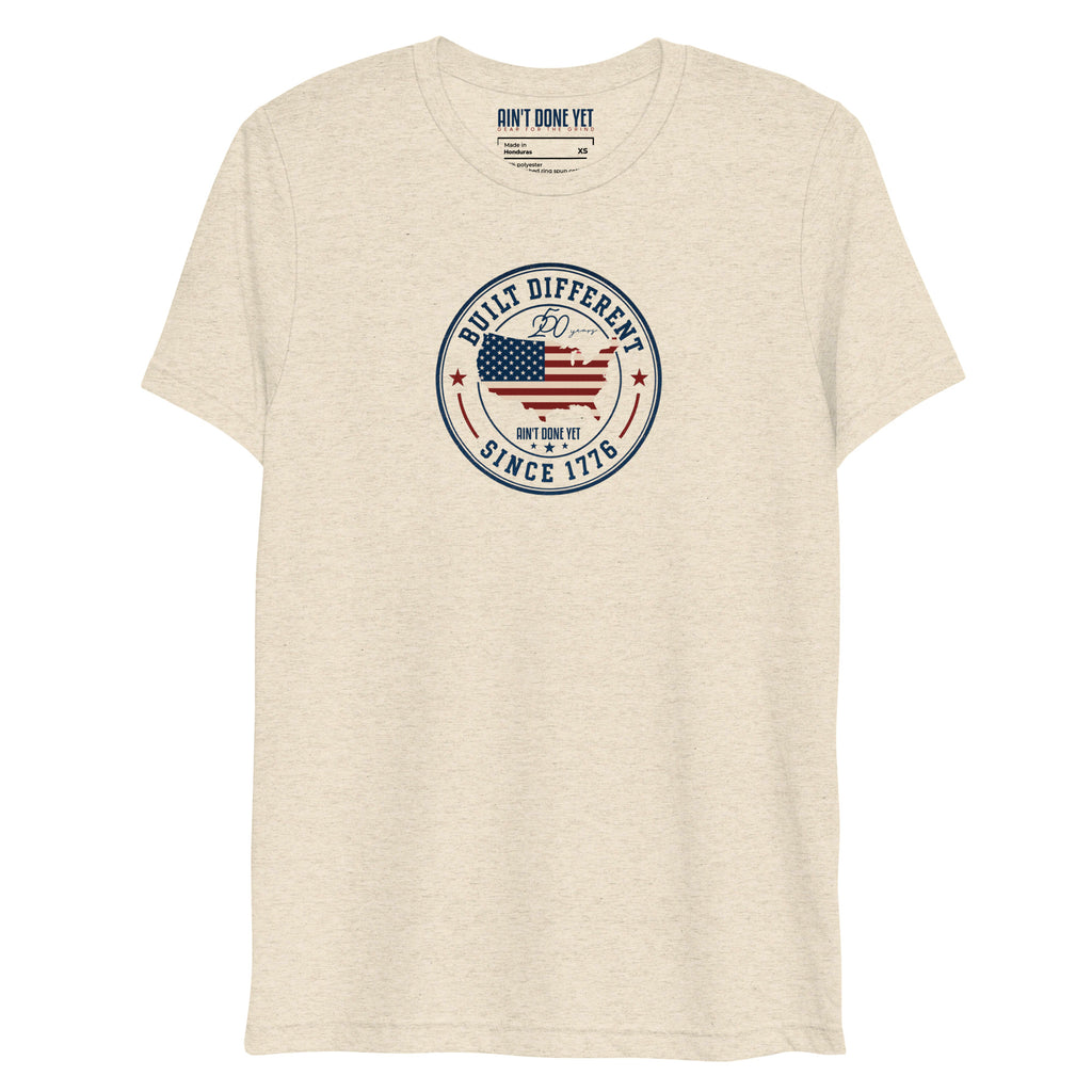 Built Different America 250 oatmeal tri-blend t-shirt featuring patriotic circular emblem with American flag and 1776 design.