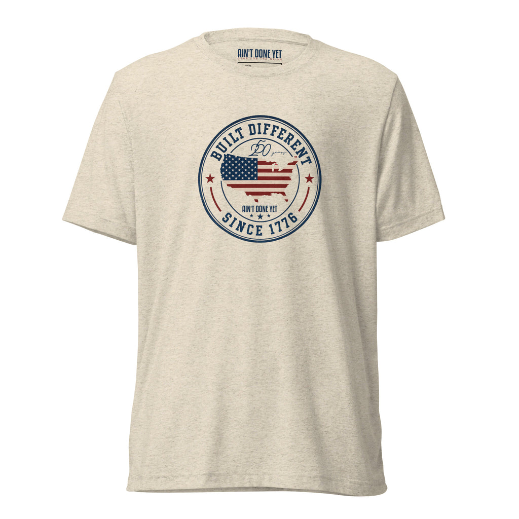 Built Different America 250 oatmeal tri-blend t-shirt featuring patriotic circular emblem with American flag and 1776 design.