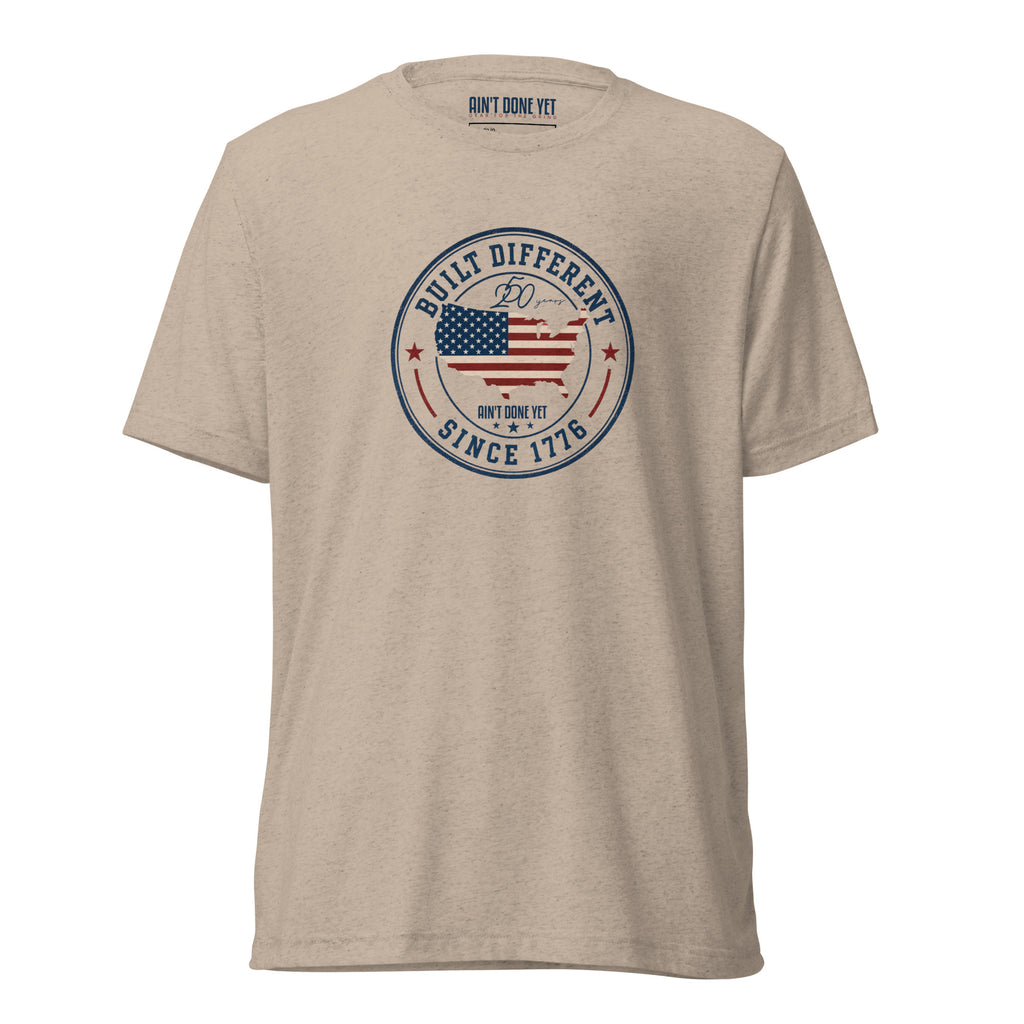 Built Different America 250 tan tri-blend t-shirt featuring patriotic circular emblem with American flag and 1776 design.